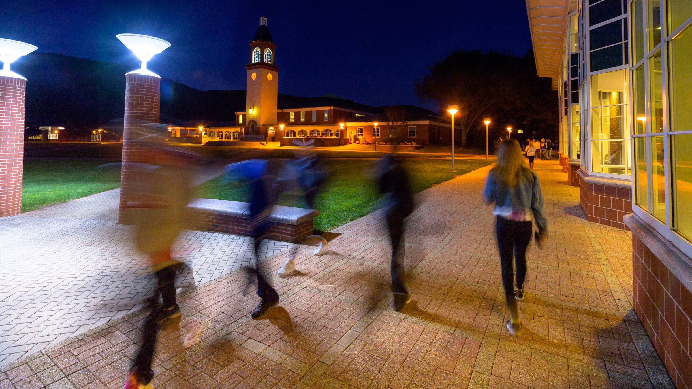 Half a dozen students walk next to the student center in with windows aglow in the evening.
